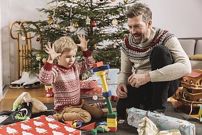 Vater und Sohn sitzen vor Geschenkeberg neben dem Christbaum und schauen lachend zu, wie ein Turm aus Bauklötzen einstürzt