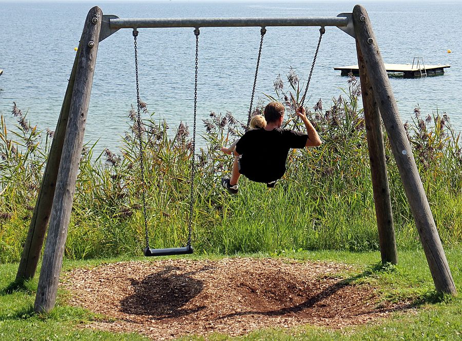 Vater mit Kind auf dem Schoß schaukelt auf einer Schaukel am See