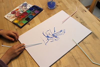 Papier mit blauem Muster aus Wasserfarben, dazu eine Hand mit Strohälmen, Wasserfabkasten und ein Glas