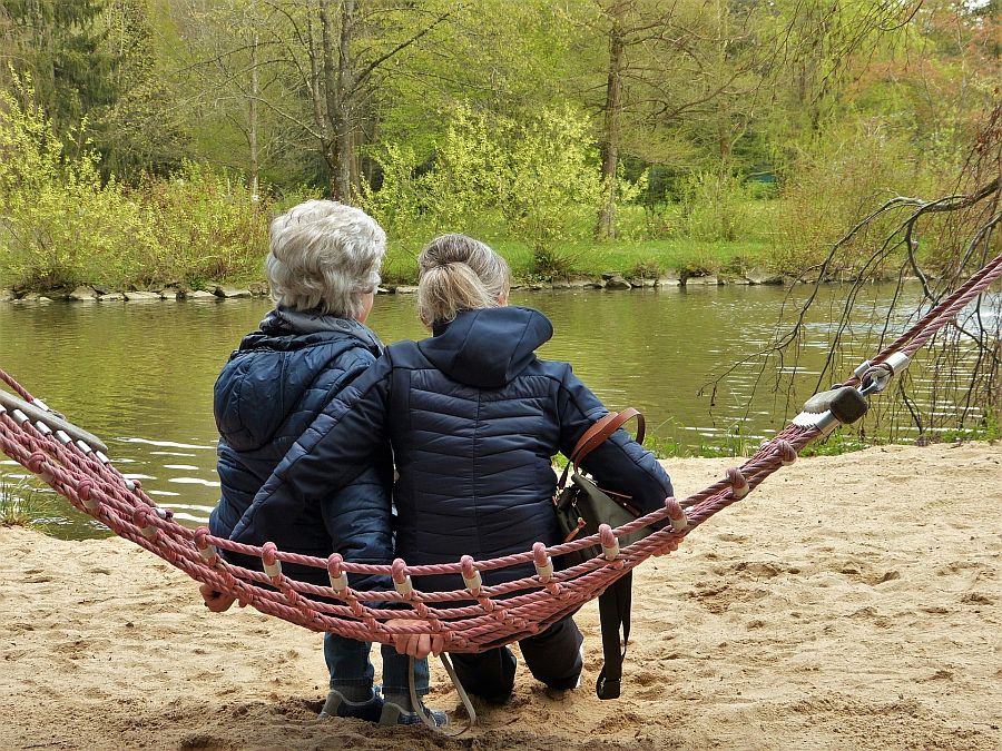 Eine ältere und eine jüngere Frauen sitzen mit dem Rücken zum Betrachter auf einer Hängematte an einem See