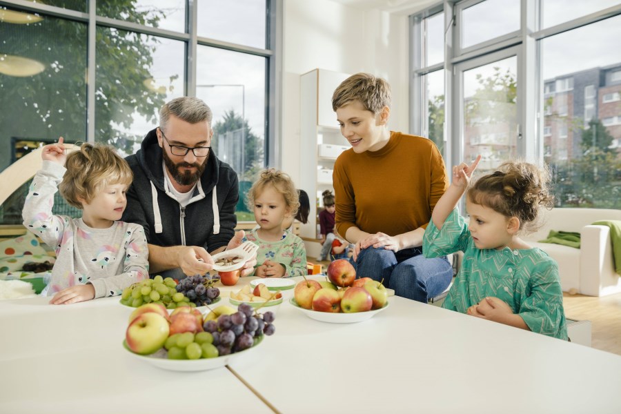 Erzieher und Erzieherin bereiten in der Kita mit Kindern frischen Obstteller vor