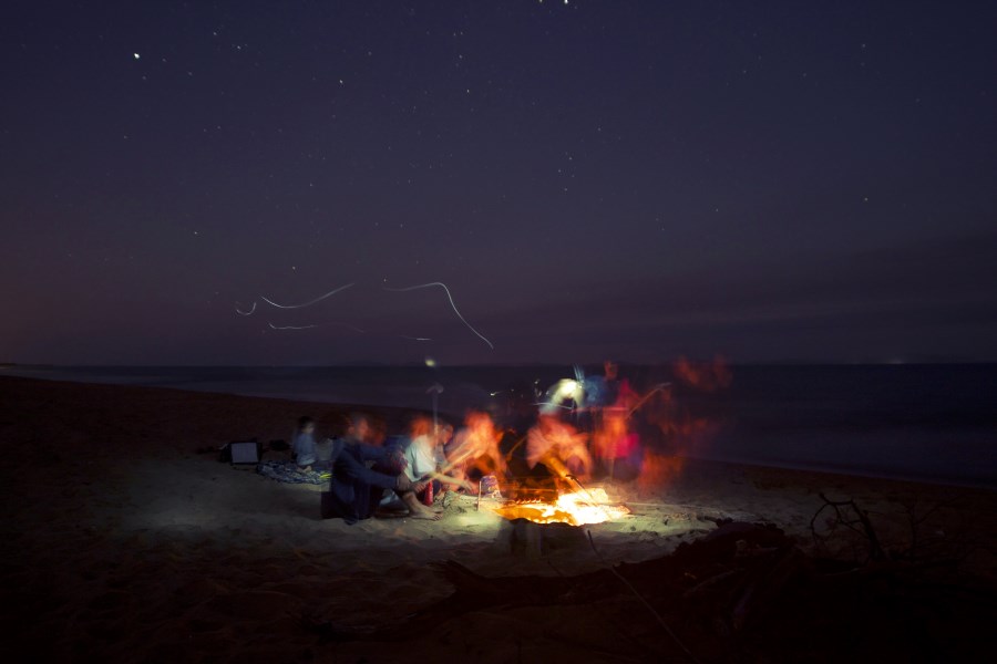 Gruppe Menschen sitzt um ein Lagerfeuer an einem Ufer am Abend