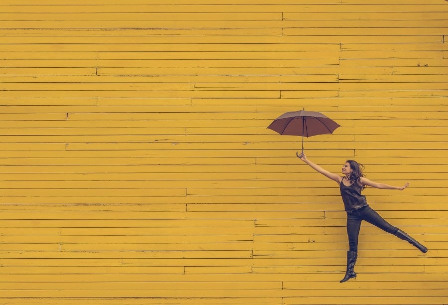 Frau fliegt mit Regenschirm vor gelber Wand