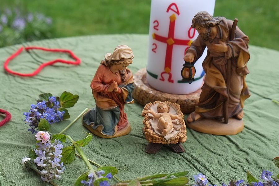 Maria mit Joseph und Kind in Krippe vor Osterkerze, rundherum Blumen