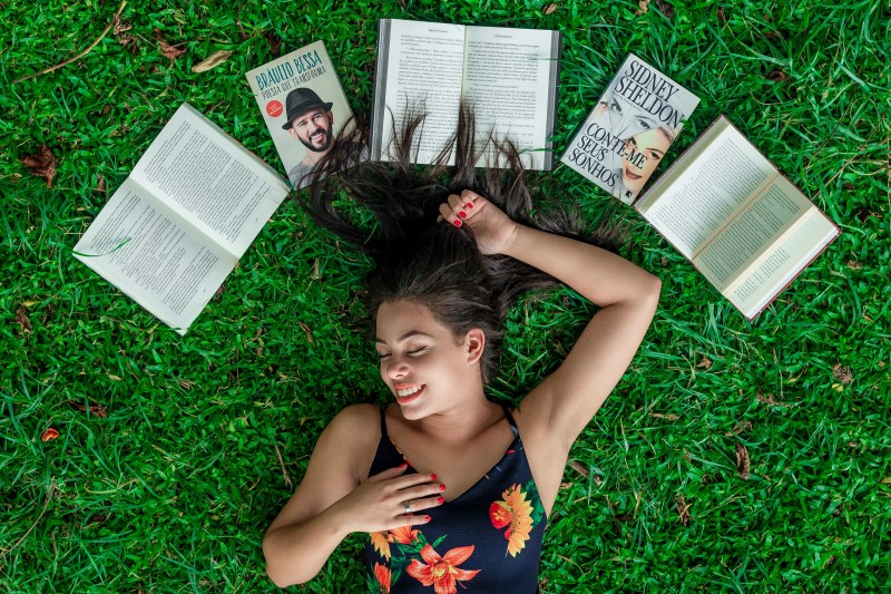 Eine Frau liegt lachend auf einer Wiese, umgeben von Büchern