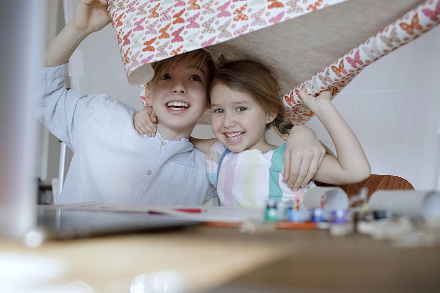 zwei Kinder halten lachend Bogen Geschenkpapier über sich