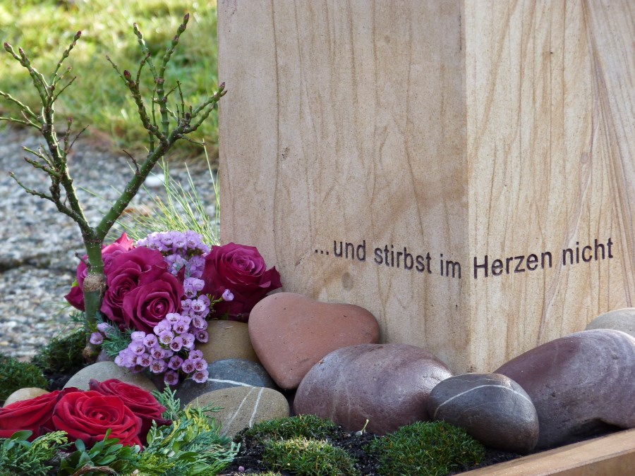 Grabstein mit der Inschrift "...und stirbt im Herzen nicht" mit Steinen und Blumen davor