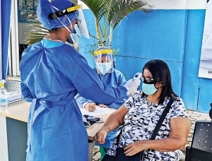 Auf dem Foto sieht man eine medizinische Angesellte. Sie trägt eine Schutzausrüstung und misst bei einer Frau Fieber. Beide tragen eine Maske.