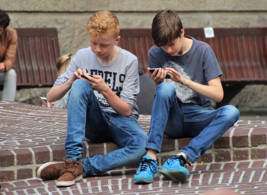 Zwei Jungen sitzen auf einem Pflastervorsprung und wischen mit den Fingern über ihre Smartphone-Screens.