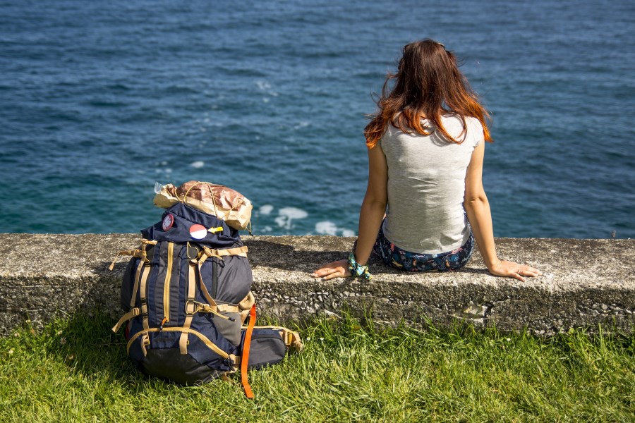 Auf dem Foto sieht man eine Frau von hinten am Meer. Hinter ihr steht ein Wanderrucksack