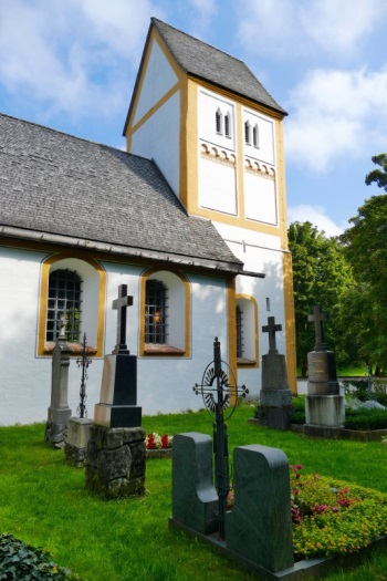 Im Vordergrund sind Gräber des Friedhofes von Heilig Kreuz zu sehen, im Hintergrund ist der Turm von Heilig kreuz im Fokus.