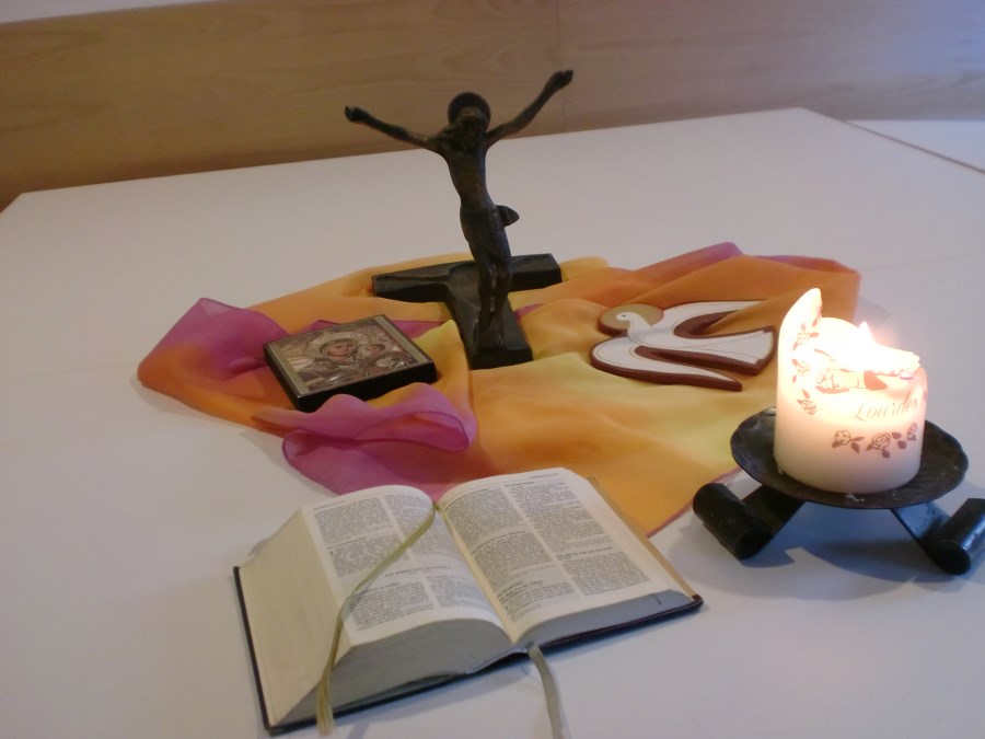 In der Tischmitte liegen ein Kreuz, eine Ikone, eine Taube, eine brennende Kerze und eine aufgeschlagene Bibel.