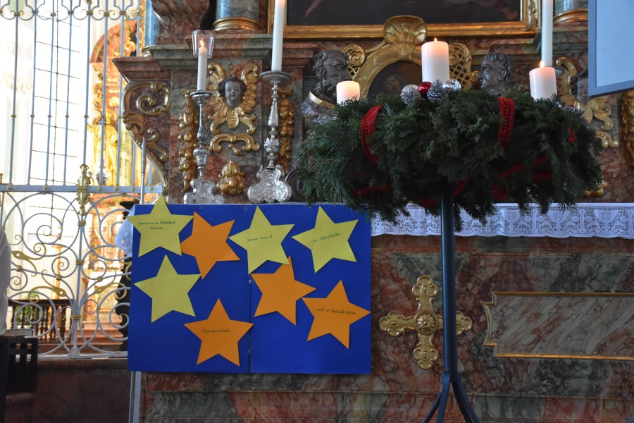 Neben dem brennenden Adventskranz hängen auf blauem Hintergrund gelbe Sterne.