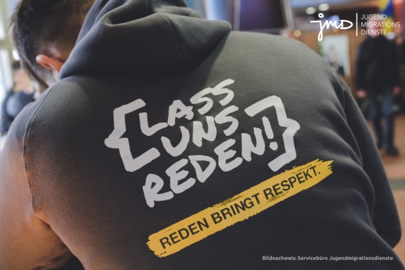 Auf dem Foto ist ein junger Mann von hinten zu sehen, der einen Pullover trägt, auf dem der Schriftzug "Lass uns reden. Reden bringt Respekt" steht.