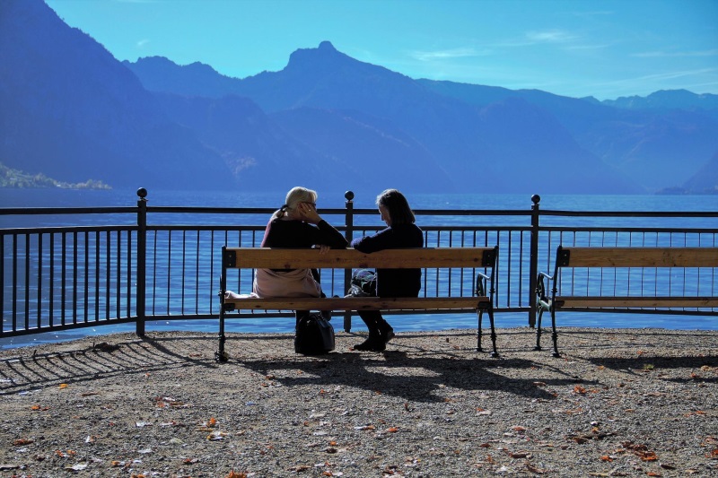 Auf dem Foto sind zwei Frauen zu sehen, die an einem Seeufer auf einer Bank sitzen und sich unterhalten.