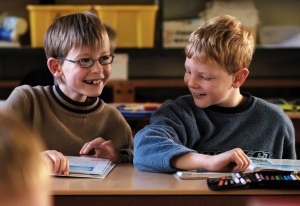 Auf dem Foto sind zwei Schulkinder zu sehen, die nebeneinander an einer Schulbank sitzen.