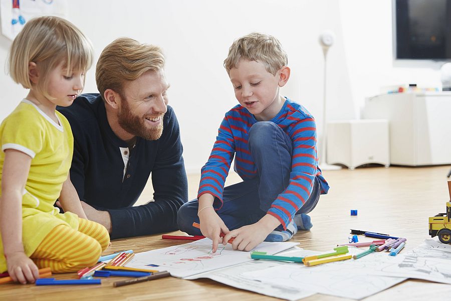 Vater und Kinder liegen fröhlich mit Malbüchern und Stiften auf dem Wohnzimmerfußboden