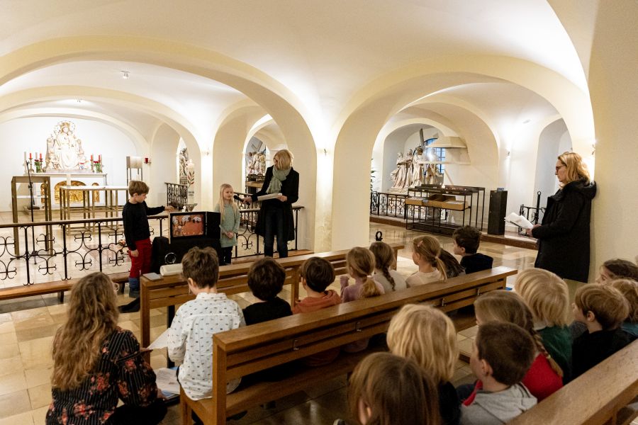Eine Schülerin und ein Schüler stellen ihren Klassenkameradinnen der Klasse 1b der Franziskus-Grundschule im Bürgersaal der Unterkirche ein Kunstwerk vor