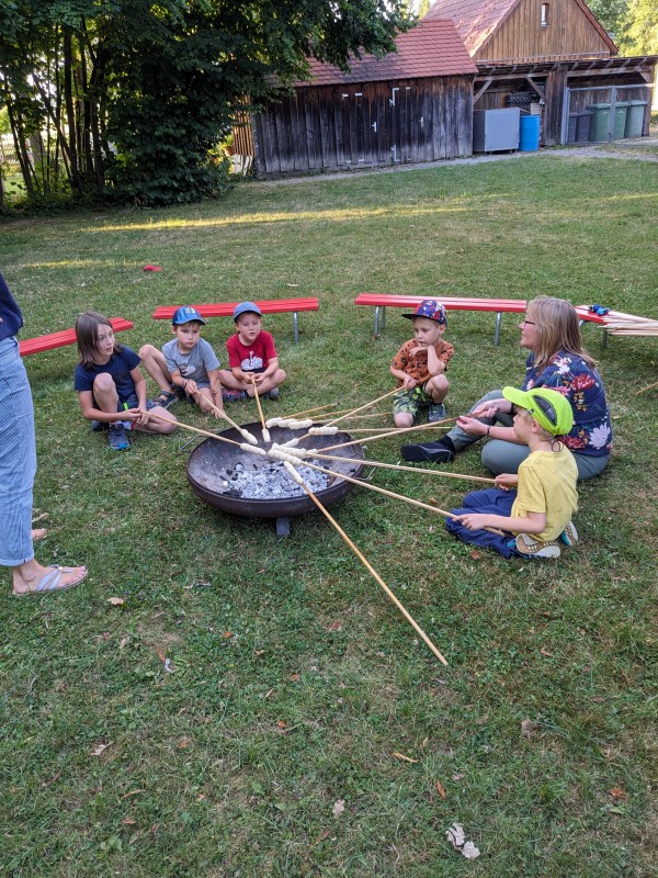 Kinder hocken im Gras um eine Feuerschale und halten Stockbrot über die Glut