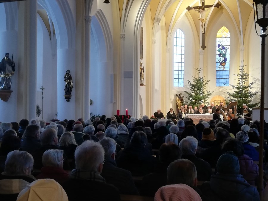 Gefüllte Kirche in Richtung Altarraum, der hell erleuchtet ist - dort stehen Sänger und Instrumentalisten, u.a. eine Harfe
