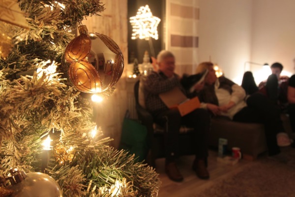 Auf dem Foto ist ein geschmückter Weihnachtsbaum mit Lichterkette zu sehen. Im Hintergrund sitzt eine Familie auf dem Sofa.