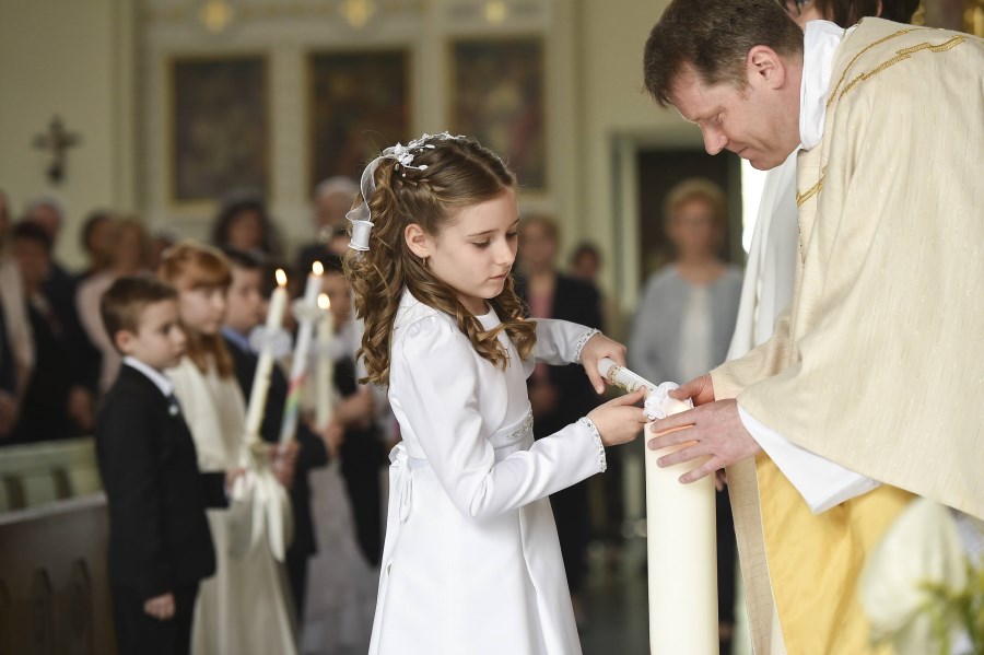 Auf dem Foto ist ein Mädchen zu sehen, das in einer Kirche gemeinsam mit einem Pfarrer ihre Kommunionkerze anzündet.
