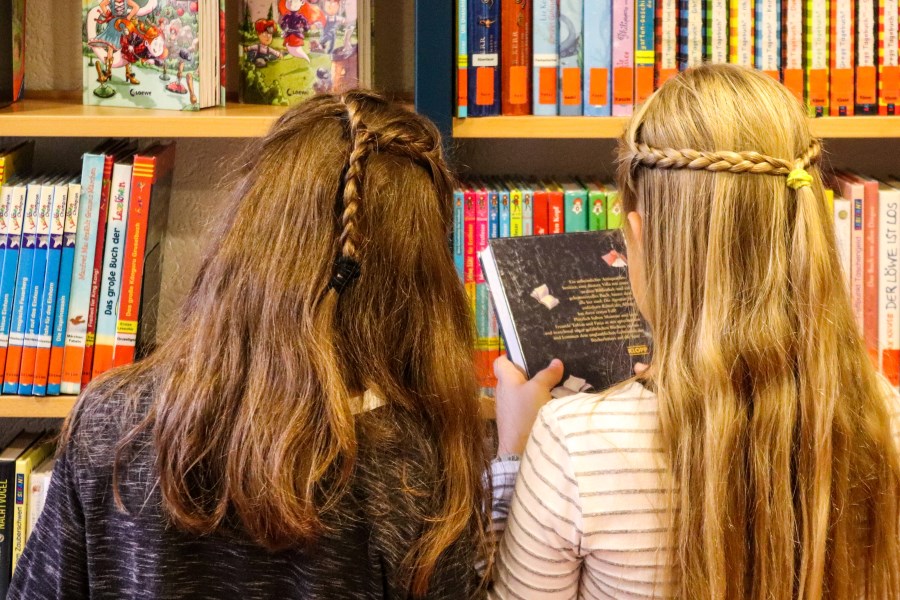 Auf dem Foto sind zwei Mädchen von hinten vor einem Regal mit Büchern in einer Bücherei zu sehen.