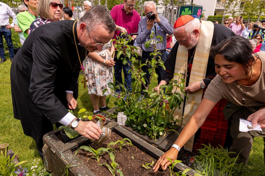 Landesbischof Christian Kopp, Kardinal Reinhard Marx und die Künstlerin Claudia Starkloff säen Pflanzensamen in Munitionskisten auf der Landesgartenschau