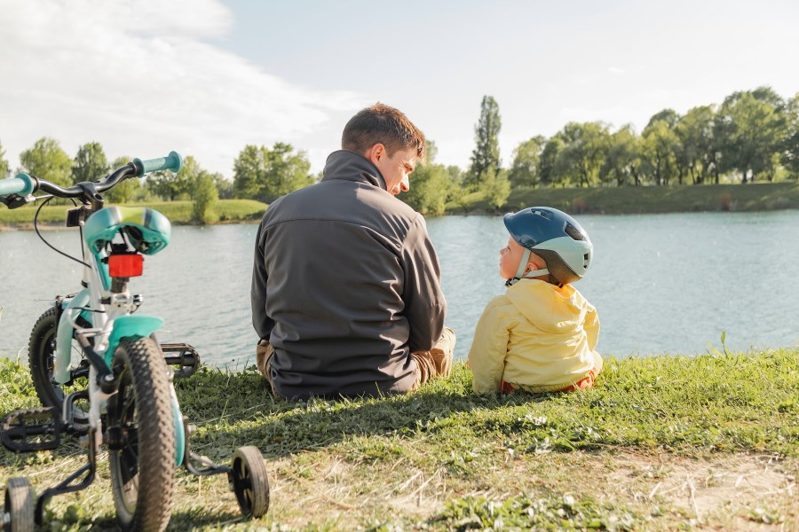 Auf dem Foto ist ein Vater zu sehen. Er sitzt mit seinem kleinen Sohn an einem Fluss und unterhält sich. Der Sohn trägt einen Fahrradhelm. Hinter den beiden liegt ein Laufrad