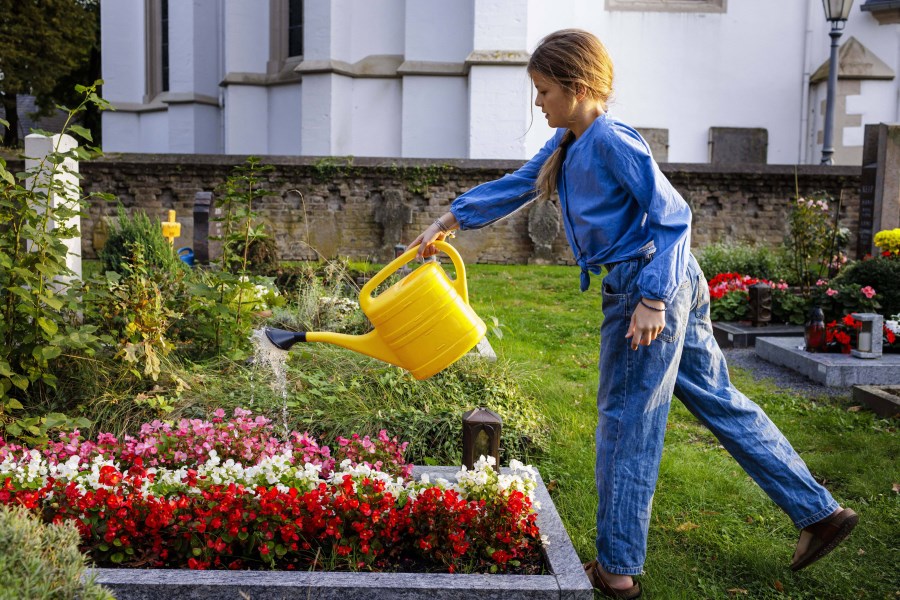 Auf dem Foto ist ein Mädchen auf einem Friedhof zu sehen. Das Mädchen gießt mit einer Gießkanne Blumen auf einem Grab.
