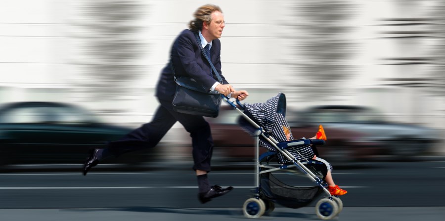 Auf dem Foto ist ein Mann im Anzug zu sehen. Er rennt und schiebt einen Kinderwagen vor sich mit einem Kleinkind darin