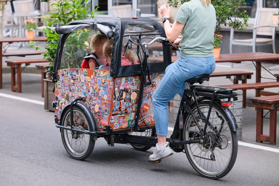 Auf dem Foto ist eine Mutter zu sehen, die mit einem Lastenfahrrad fährt. Im Lastenfahrrad sitzen Kinder.