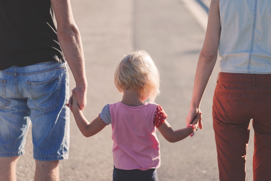 Auf dem Foto sind ein Vater und eine Mutter von hinten zu sehen. In der Mitte an den Händen von beiden Läuft ein Kleinkind.
