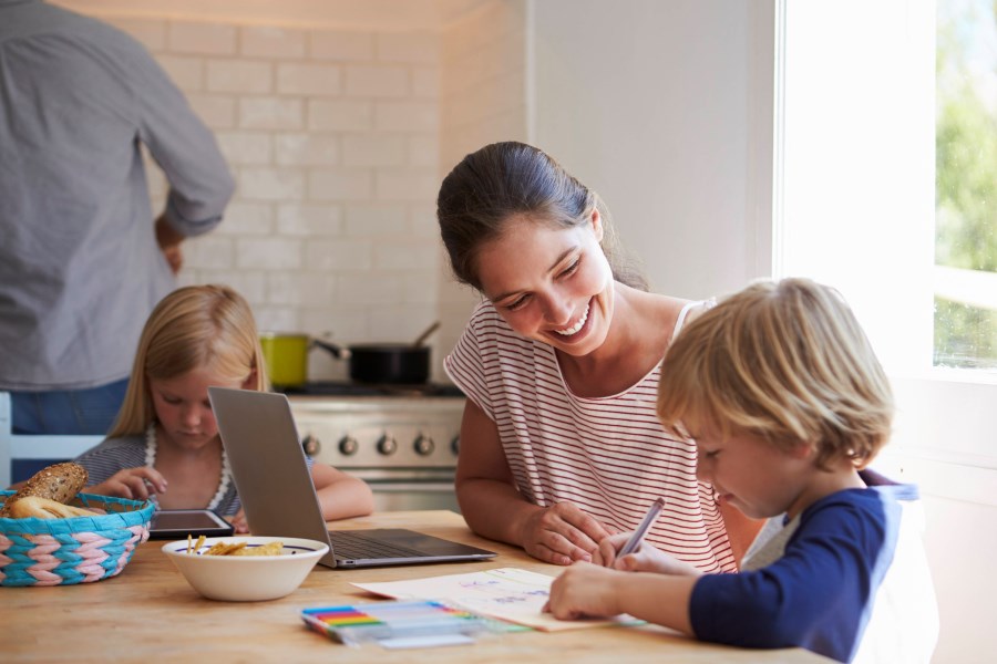 Auf dem Foto ist eine Mutter zu sehen. Sie sitzt am Küchentisch. Neben ihr sitzt ein Junge, der Vorschulübungen macht. Sie lächelt ihm zu.