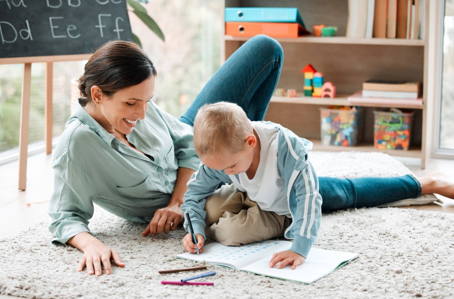 Auf dem Foto ist eine Mutter zu sehen. Sie liegt im Kinderzimmer auf dem Boden und lächelt ihr Kind an. Das Kind macht gerade Vorschulübungen.