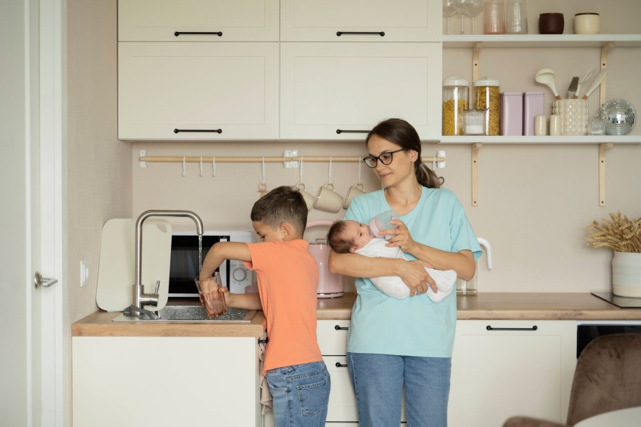 Auf dem Foto ist eine Mutter mit ihren Kindern zu sehen. Sie steht in der Küche und hält ein Baby im Arm. Der Sohn schenkt sich ein Glas Wasser ein. <br/>Alleinerziehend