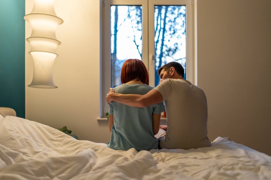 Auf dem Foto ist ein Paar zu sehen. Es sitzt auf dem Bett, mit dem Rücken zur Kamera. Der Mann umarmt und tröstet die Frau.