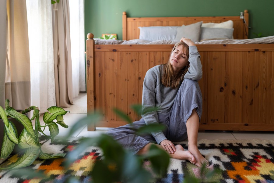 Auf dem Foto ist eine Frau zu sehen. Sie ist traurig und sitzt vor einem Bett auf dem Boden.