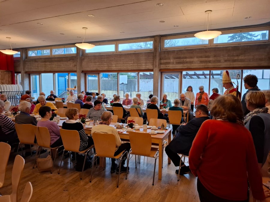 Pfarrsaal. An zwei langen Tischreihen sitzen Damen und Herren bei Kaffee und Kuchen und hören dem Nikolaus zu.