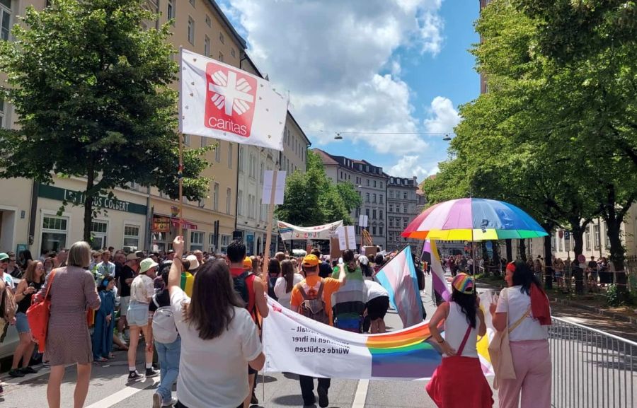 Demonstranten mit Caritas-Fahne und Schirm in Regenbogenfarben auf der Straße in einer Stadt im Sommer