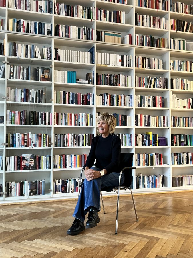 Ulrike Kriener sitzt lachend auf einem Stuhl vor einem Bücherregal