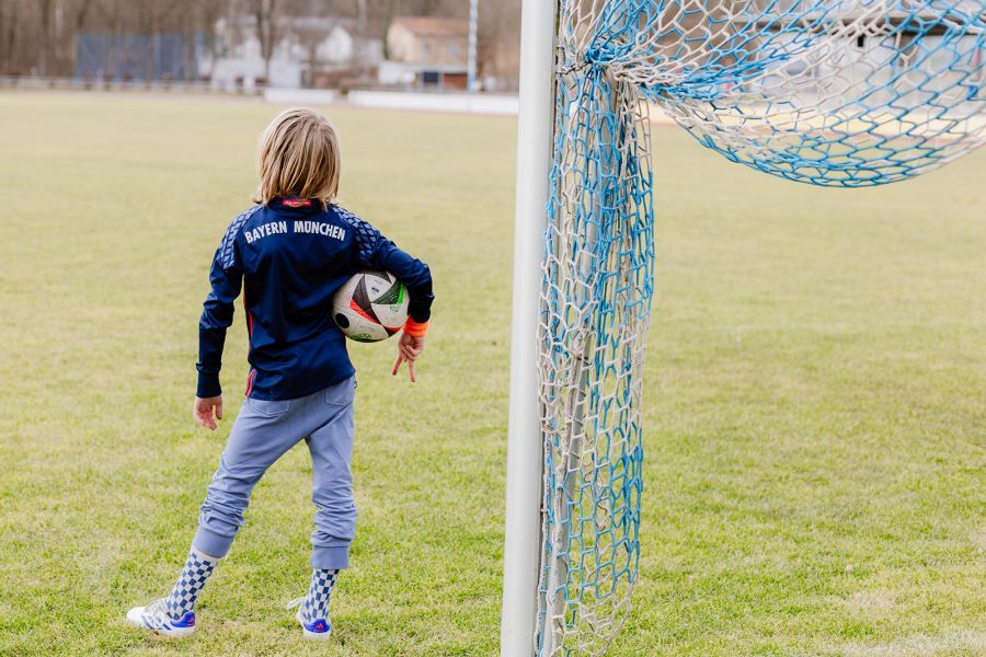 Ein Junge im Fußballtrikot steht mit einem Ball unter dem Arm mit dem Rücken zur Kamera neben einem Tor auf einem Fußballplatz