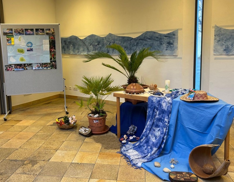 Dekoration zum Weltgebetstag 2025 im Pfarrsaal St. Hildegard. Palmen, an der Wand gezeichnete Berge, blaue Tücher als Wasser.