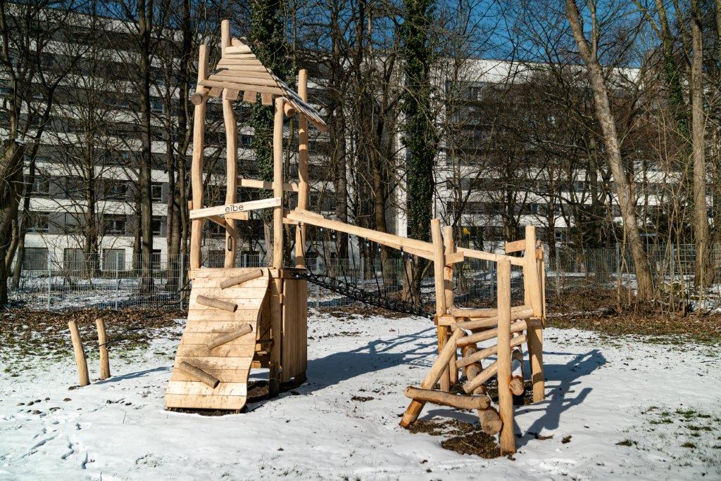 Kletterturm aus Holz mit Seilhängebrücke im verschneiten Garten des Kinderhauses Maria Schutz, im Hintergrund die Heliosklinik Pasing.