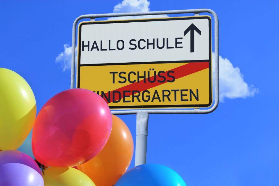 Verkehrsschild mit einem durchgestrichenen "Tschüß Kindergarten" und einem "Hallo Schule" hinter bunten Luftballons