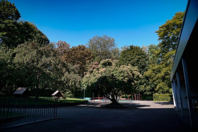 Blick in den sommerlichen Garten der Kita Maria Schutz mit vielen Laubbäumen, dazwischen Spiel- und Klettergeräte