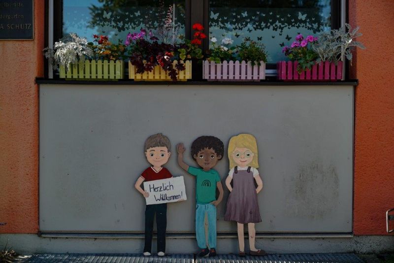 Drei gemalte Kinderfiguren mit einem Schild "herzlich willkommen" vor dem Eingang zur Kita Maria Schutz