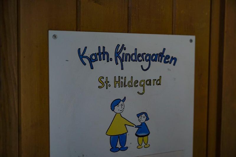 Schild am Eingang zur Kita mit der Aufschrift: Kath. Kindergarten St. Hildegard" und zwei gezeichneten Figuren in blau und gelb