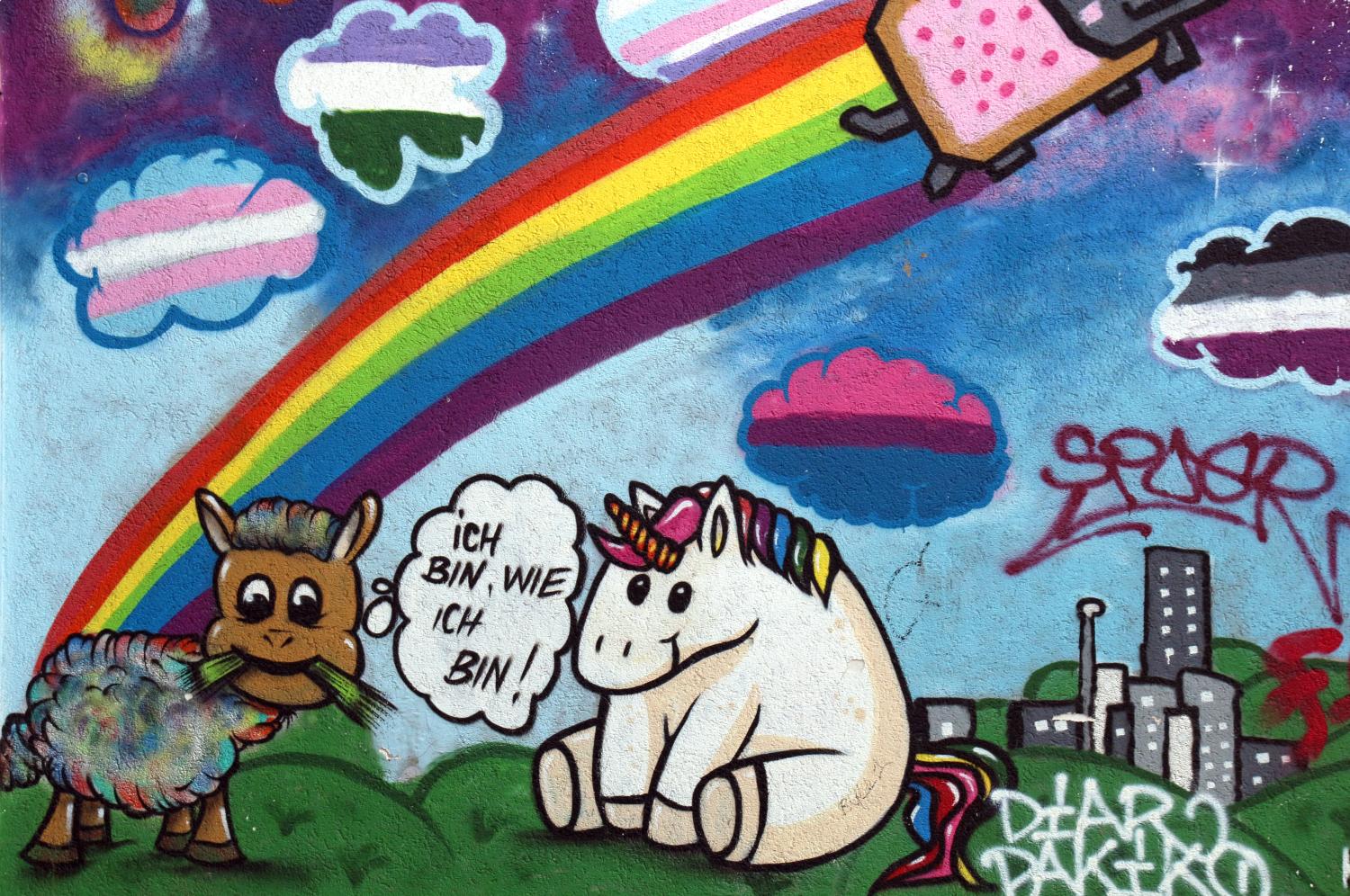 Graffito<br/>ein Regenbogen, darüber unter anderem trans Farben, darunter eine Wiese, ein Pferd mit buntem Fell grast, eine Sprechblase bei ihm: ich bin, wie ich bin, daneben ein weisses Einhorn mit rosa