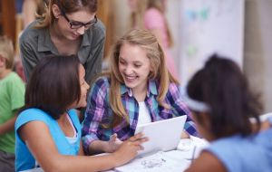 Zwei sitzende und eine stehende Schülerinnen unterhalten sich lachend vor einem Tablet-Computer.